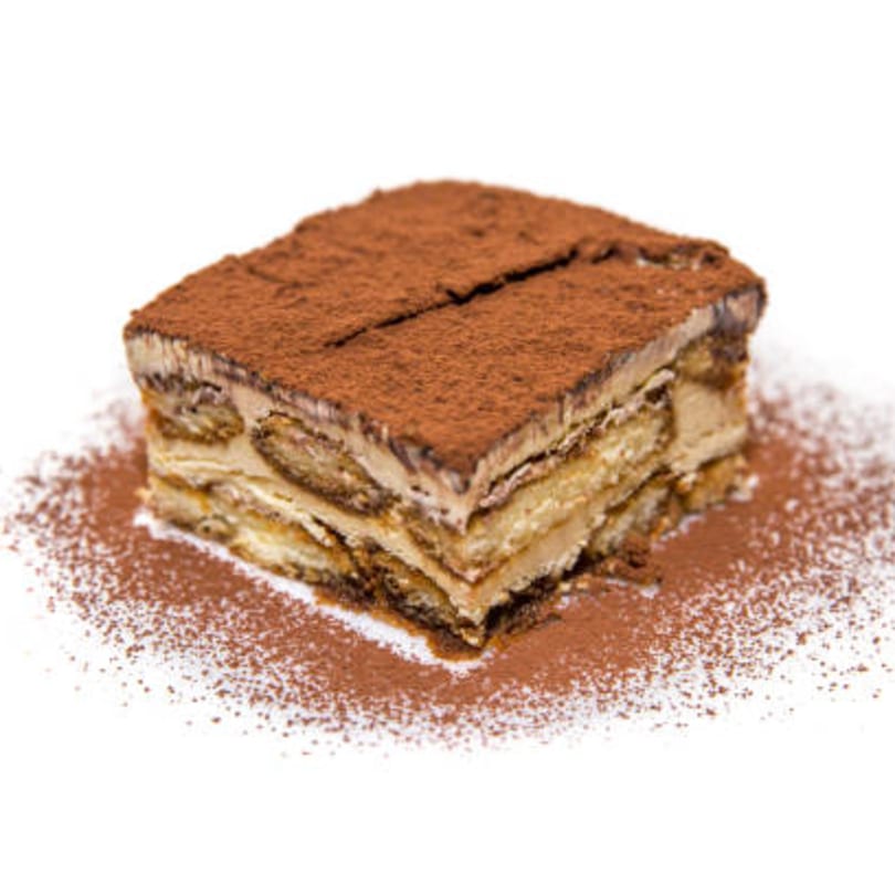 A square slice of tiramisu with cocoa powder on top and scattered around on a white background.